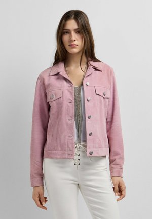 Junge Frau trägt eine hellrosa Jacke mit Knöpfen, weiße Schnürhose und eine lange silberne Kette, steht vor einem einfarbigen grauen Hintergrund.