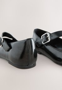 Schwarze Mary Jane Lacklederschuhe mit silbernen Schnallenriemen, flachen Sohlen, auf neutralem Hintergrund gezeigt.