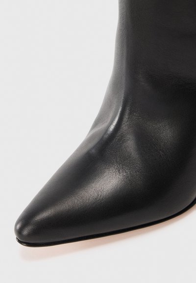Gros plan sur une botte en cuir noir à bout pointu, avec une texture lisse et un design élégant sur un fond clair.