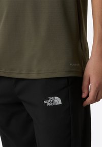 Olivno zelena srajca z teksturirano tkanino, z oznako "FLASHDRY" blizu roba. Kombinirana s črnimi hlačami z belim logotipom.