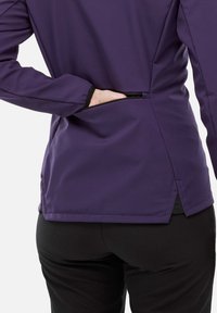 Personne portant une veste violette et un pantalon noir avec une main dans une poche zippée au bas du dos de la veste.