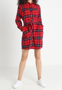 Abito camicia a quadri rosso e navy con maniche lunghe, chiusura a bottoni, due tasche laterali e cintura in vita, realizzato in tessuto morbido. Indossato con sneaker bianche.