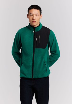 Man wearing green and black zip-up fleece jacket with hands behind back, standing against plain light background.