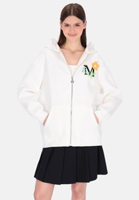 Fehér cipzáras kapucnis pulóver elülső zsebekkel, kapucnival és pasztell színű virágos hímzéssel. Fekete pliszírozott szoknyával párosítva.