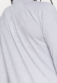 Vaaleanharmaa urheilupaita, jossa on teksturoitu pinta. Sisältää olkaimen sauman ja pieniä heijastavia yksityiskohtia selkäpuolella.