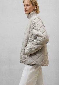 Helle beige gesteppte Jacke mit hohem, geripptem Kragen und seitlichen Taschen. Sie hat einen lässigen Schnitt und eine weiche, glatte Textur. Kombiniert mit weißen Hosen.