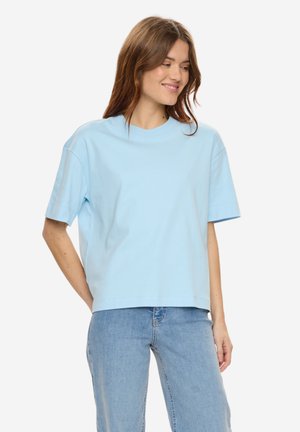 Frau mit hellbraunen Haaren, die ein lockeres hellblaues T-Shirt und blaue Jeans trägt, lächelt und schaut nach links vor einem schlichten Hintergrund.