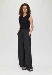 Zwart mouwloos shirt met ruches van kant, gecombineerd met losse zwarte palazzo-pantalons. Het model heeft krullend haar en staat met handen in de zakken.
