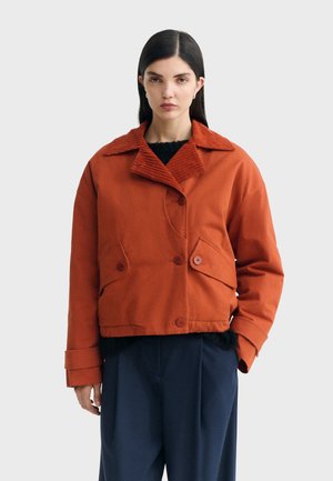 Femme aux cheveux raides et foncés porte une veste boutonnée orange rouille avec un grand col et un pantalon large bleu marine, debout sur un fond gris.