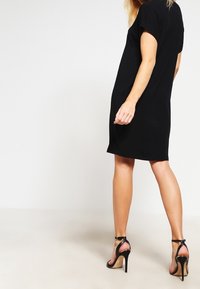 Černé šaty s krátkým rukávem a volným střihem, vyrobené z hladké látky. Na sobě má černé sandály na vysokém podpatku se straps, zobrazeny ze zadního pohledu.