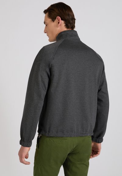 Uomo che indossa una giacca a maniche lunghe grigio scuro e pantaloni verde oliva di spalle su uno sfondo semplice.