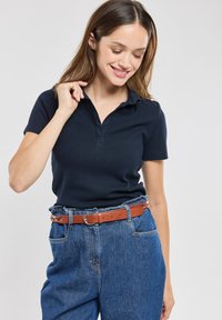 Polo shirt blu navy con maniche corte e colletto a 3 bottoni, abbinato a pantaloni in denim a vita alta e una cintura in pelle marrone.