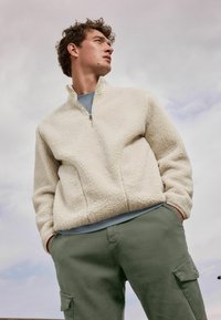 Pull en polaire beige clair avec col haut, détail zippé et texture douce, assorti à un pantalon cargo vert olive avec poches latérales.