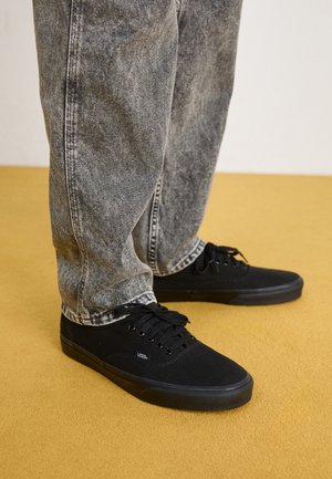 Personne portant un jean gris délavé et des baskets Vans noires debout sur un sol jaune moutarde.