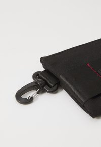 Zwarte stoffen pouch met een gestructureerd oppervlak, voorzien van een zwarte metalen clip en details in rode stiksels. Compact en rechthoekig ontwerp.
