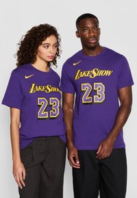 Fialové bavlněné trička s krátkým rukávem, s potiskem "LakeShow" a číslem "23" v žluté a černé barvě. Logo Nike na levém horním rohu.