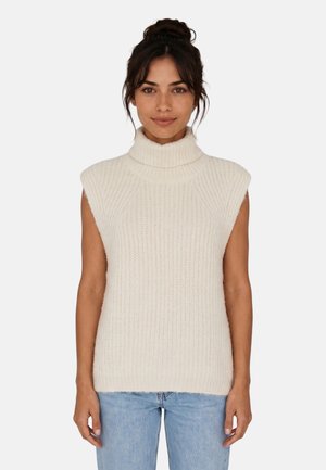 Giovane donna con capelli scuri raccolti in uno chignon, che indossa un maglione a collo alto senza maniche color crema e jeans azzurri, in piedi davanti a uno sfondo semplice.