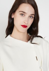 Top blanco estructurado con un escote asimétrico y detalles de hardware dorado. Textura suave, mangas largas y un corte relajado.