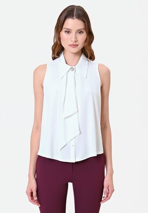 Jeune femme aux cheveux bruns mi-longs, portant un chemisier blanc sans manches avec un détail volanté et un pantalon violet foncé, face à l'avant.