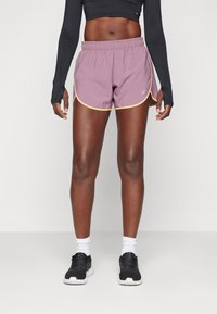 Violet sportiniai šortai su elastinga juosta, kontrastinga geltona apdaila ir logotipo detalė. Modelis dėvi juodas sportines batus su baltais sockais.