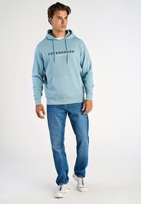 Sudadera azul claro con cordones blancos y bolsillo frontal, que presenta el texto "Copenhagen Lindbergh". Combinada con jeans azules y zapatillas blancas.