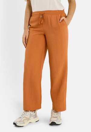 Femme portant un pantalon large orange rouille avec un cordon à pompons et des baskets blanches, debout avec une main dans la poche.