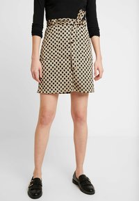 Falda beige con lunares negros, cuenta con una cintura atada, dos bolsillos frontales y un corte recto. Llevada con mocasines negros.