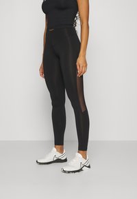 Magas derekú fekete leggings, sima textúrával és átlátszó hálós panelekel az oldalán, fehér sportcipővel párosítva, fekete részletekkel.