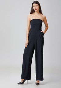 Combinaison noire sans bretelles avec un buste ajusté, des pantalons larges et des poches latérales. Fabriquée en tissu lisse, associée à des talons noirs.