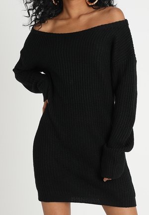 Femme portant une robe pull en maille noire épaules dénudées, debout avec une main sur la hanche et les cheveux bouclés lâchés, sur un fond uni.