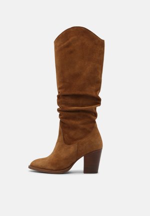 Botas castanhas de camurça até ao joelho, com cano solto, bico fino e salto empilhado. Textura suave e design minimalista.