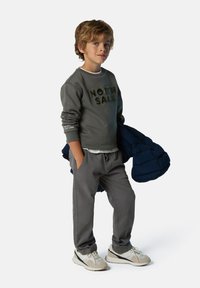 Felpa grigia con la scritta "NORTH SAILS" in lettere scure, pantaloni sportivi grigi e sneaker beige. Tiene in mano un giubbotto imbottito blu.