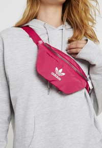Rosa Adidas midjeväska med zip-stängning, som har en vit logo, justerbar rem och en slät, glänsande tygtextur.
