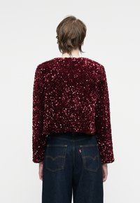 Izgriezta bordo jaka, pārklāta ar pērlītēm, ar garām piedurknēm un apaļu kakla izgriezumu, apvienota ar tumši zilu džinsu biksēm, kas ir ar aizmugurējām kabatām.