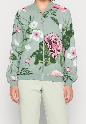 Vrouw draagt een lichtgroen jasje met rits met grote roze en witte bloemenprints en lichtbeige broek.