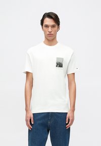 Camiseta blanca de algodón con un estampado gráfico de un bolsillo que representa un horizonte urbano, cuello redondo y mangas cortas. Unos pantalones vaqueros de mezclilla complementan el conjunto.