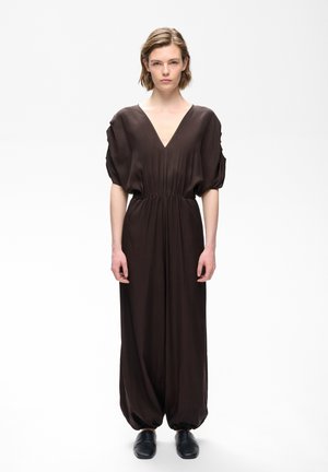 Person står iført en løs, mørkebrun jumpsuit med V-hals, samlet kant og sorte sko mod en ensfarvet hvid baggrund.