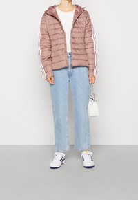 Jasnoróżowa kurtka puchowa z kapturem, z białymi paskami po bokach, w zestawie z jasnoniebieskimi dżinsami i białymi sneakersami. Mała biała torebka jako dodatek.