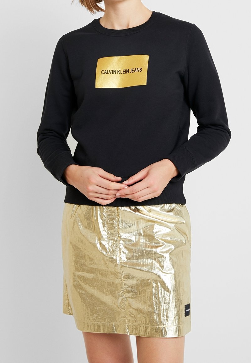 Sudadera negra con un logotipo rectangular dorado que presenta "CALVIN KLEIN JEANS". Combinada con una minifalda metálica dorada brillante.