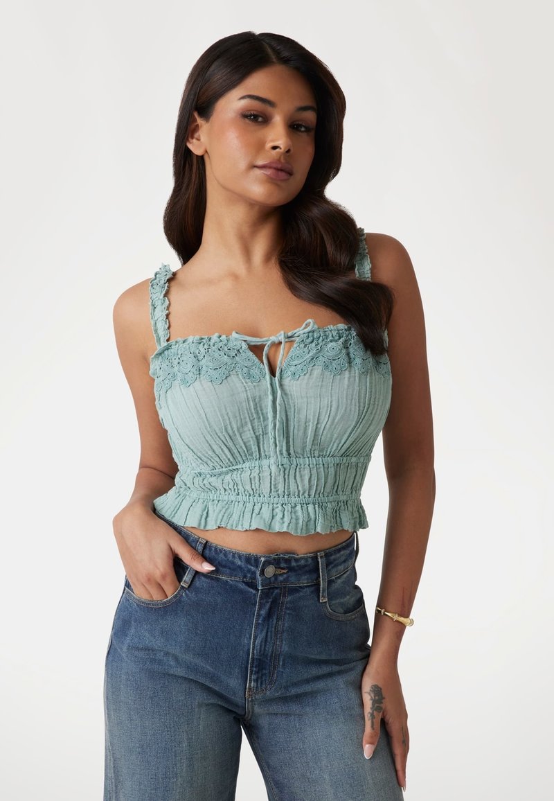 Light teal cropped top with lace trim and adjustable tie detail. Features a textured body and ruffled hem, paired with high-waisted blue jeans.