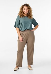 Top teal leggero con maniche corte abbinato a pantaloni marroni a vita elastica e a lunghezza intera. Il modello indossa scarpe piatte beige. Sfondo neutro.