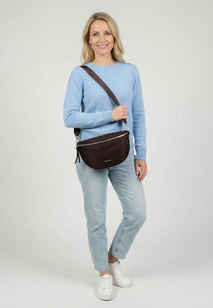 Lachende vrouw in lichtblauwe trui, lichte spijkerbroek en witte sneakers, die een bruine schoudertas met ritssluiting vasthoudt, staand tegen een effen achtergrond.