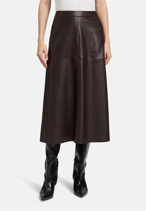 Vrouw draagt een donkerbruine leren midirok met hoge taille, gecombineerd met zwarte, kniehoge laarzen met hakken, staand tegen een witte achtergrond.