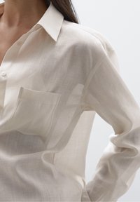 Lyse beige knap-ned skjorte lavet af gennemsigtigt stof; har en brystlomme og en afslappet pasform med klassisk krave og lange ærmer.