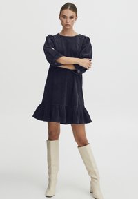 Marineblaues Cord-Minikleid mit gepufften Ärmeln, einem rüschierten Saum und Knopfdetails. Kombiniert mit kniehohen weißen Stiefeln. Glatte Textur.