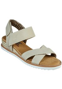 Beige sandalen met kruislings banden van gestructureerde stof; gewatteerde bruine binnenzool; witte zool met een patroon aan de binnenkant.