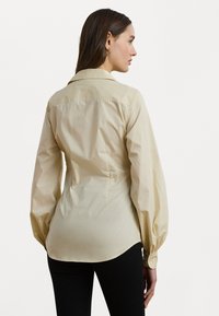 Camisa de manga longa bege com uma textura suave, punhos abotoados e um corte ligeiramente ajustado. Apresenta um colarinho padrão e um acabamento arredondado.