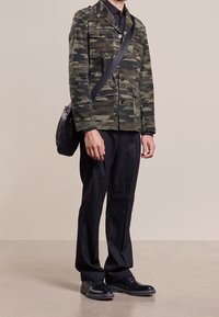 Mand iført en grøn og sort mønstret jakke, sorte bukser, sorte sko og en sort crossbody taske står på et neutralt gulv.