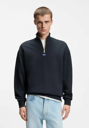 Ung mand med kort hår iført en sort sweatshirt med kvart-lynlås og lyseblå jeans, stående foran en ensartet hvid baggrund.