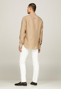 Camicia di lino marrone chiaro con chiusure a bottone, maniche lunghe e orlo curvo, abbinata a pantaloni bianchi e scarpe slip-on nere.